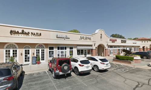 A row of storefronts with various businesses, parked cars in front, and a clear blue sky above.