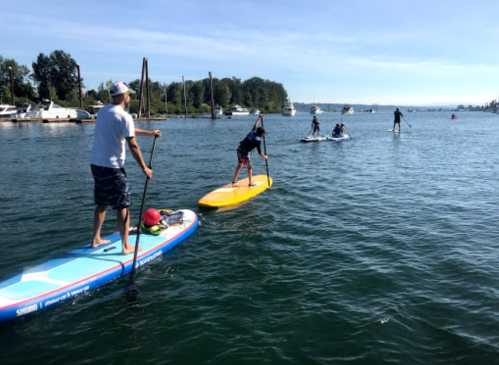 Two people paddleboarding on a calm waterway, with boats and trees in the background under a clear blue sky.