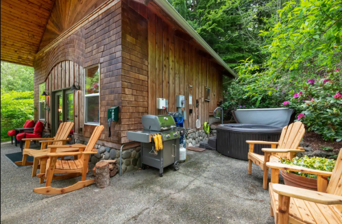 Cozy wooden cabin exterior with a grill, hot tub, and seating area surrounded by greenery and flowers.