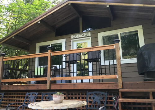 A cozy cottage with a wooden deck, two bar stools, and a round table surrounded by greenery.