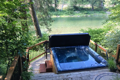 A hot tub on a wooden deck overlooking a serene river surrounded by lush greenery.