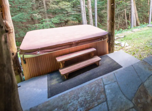 A wooden hot tub with steps, surrounded by trees and greenery, set on a stone patio.