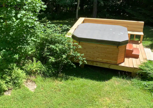 A wooden hot tub on a deck surrounded by greenery and trees in a sunny outdoor setting.