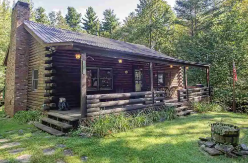 A rustic log cabin surrounded by trees, featuring a porch and a small fire pit in the front yard.