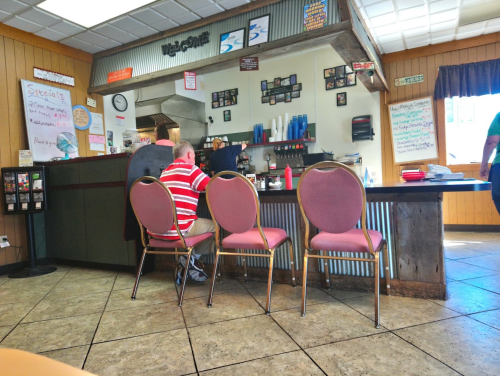 A diner interior with a man in a striped shirt sitting at the counter, chairs lined up, and a menu board in the background.