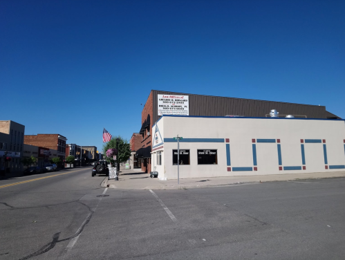 A street view of a small town with a building displaying a sign and an American flag in the background. Clear blue sky.