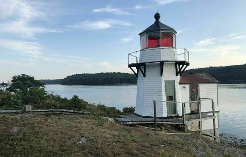 A white lighthouse with a red lantern sits by a calm river, surrounded by greenery and under a clear blue sky.