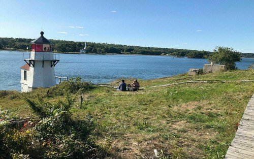 Two people sit on a grassy shore near a lighthouse, overlooking a calm river and distant trees under a clear blue sky.