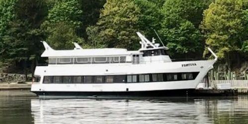 A large white boat docked on calm water, surrounded by green trees and a wooden pier.