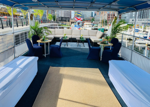 A spacious boat deck with seating, tables, and plants, overlooking a marina with boats and city buildings in the background.