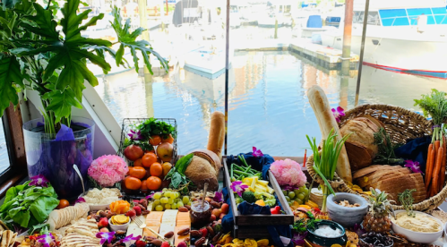 A vibrant spread of fruits, cheeses, and breads arranged beautifully by a waterfront, with boats in the background.