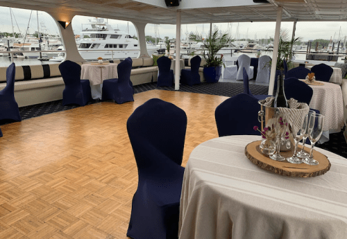 Elegant indoor event space on a boat, featuring tables, blue chair covers, and a view of yachts in the marina.
