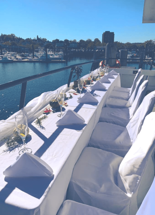 A beautifully set outdoor dining table with white linens and glasses, overlooking a marina on a sunny day.