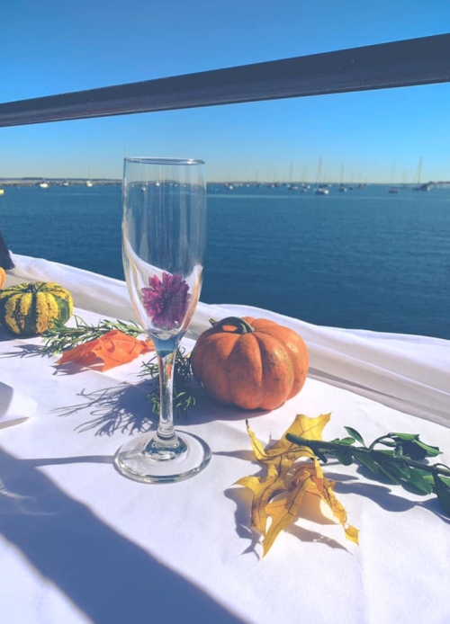 A clear glass with a flower inside, surrounded by pumpkins and leaves, with a serene waterfront view in the background.