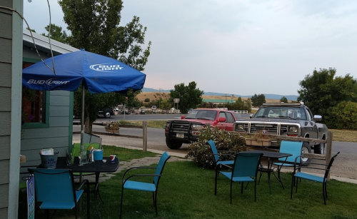 Outdoor seating area with blue chairs and a Bud Light umbrella, overlooking a road with parked trucks and trees.