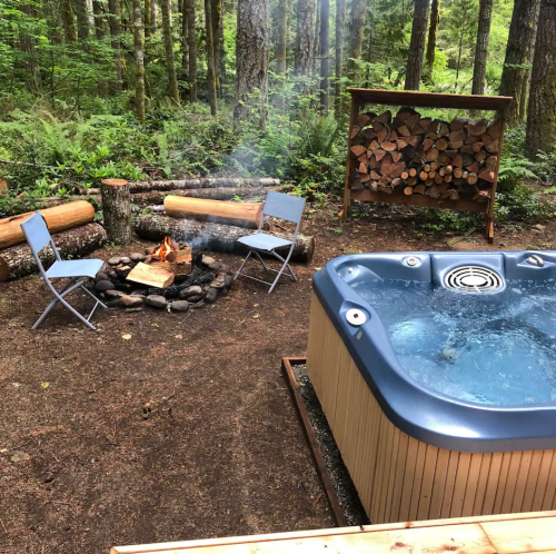 A serene outdoor setting with a hot tub, a fire pit surrounded by chairs, and stacked firewood in a forest.