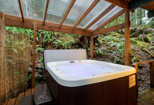 A hot tub under a wooden shelter surrounded by lush greenery and ferns in a tranquil outdoor setting.