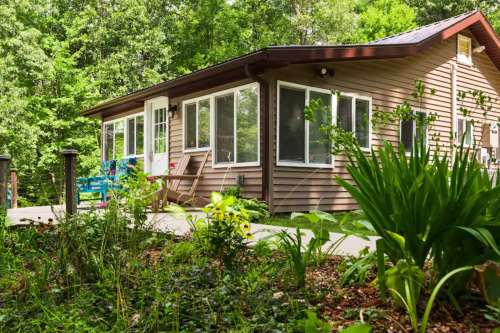 A cozy brown house surrounded by greenery and colorful flowers, with large windows and a welcoming porch.