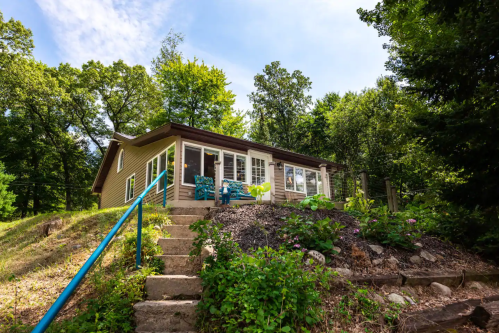A cozy cabin on a hillside, surrounded by trees, with a staircase leading up to a porch and colorful outdoor seating.