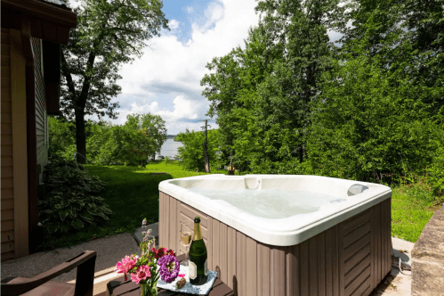 A hot tub on a patio surrounded by lush greenery, with a view of a lake and cloudy sky in the background.
