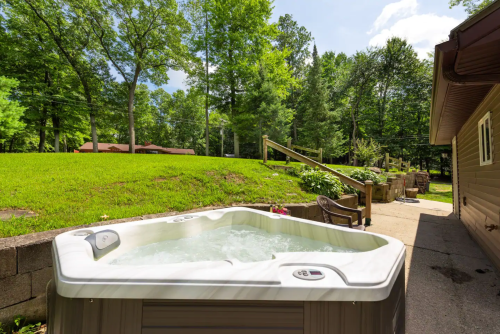 A hot tub sits on a patio surrounded by lush green trees and grass under a clear blue sky.