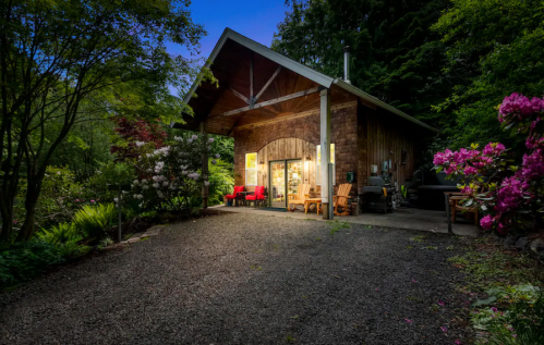 A cozy wooden cabin surrounded by lush greenery and colorful flowers, illuminated at dusk.