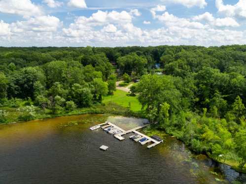 Aerial view of a serene lake with a dock, surrounded by lush green trees and a clear blue sky.