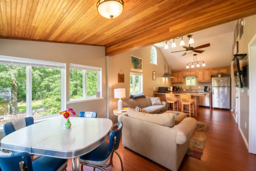 Cozy living space with a dining table, sofa, and kitchen, featuring large windows and wooden ceiling. Bright and inviting atmosphere.