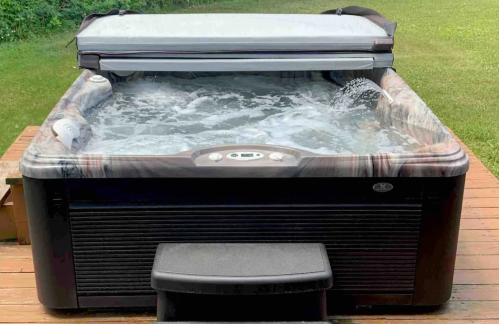 A hot tub with bubbling water, partially covered, on a wooden deck surrounded by green grass.