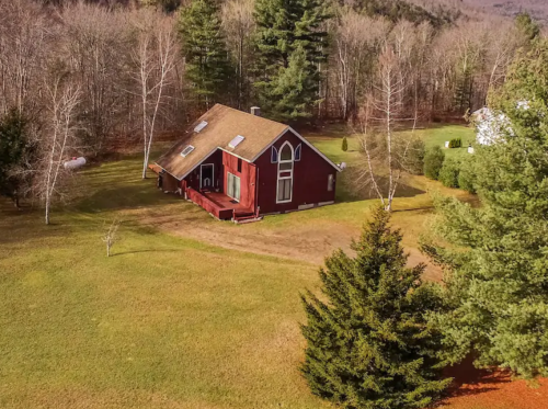 A red house with a sloped roof sits in a grassy area surrounded by trees and a wooded landscape.