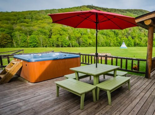 A wooden deck with a hot tub, green picnic tables, and a large red umbrella overlooking a lush green landscape.