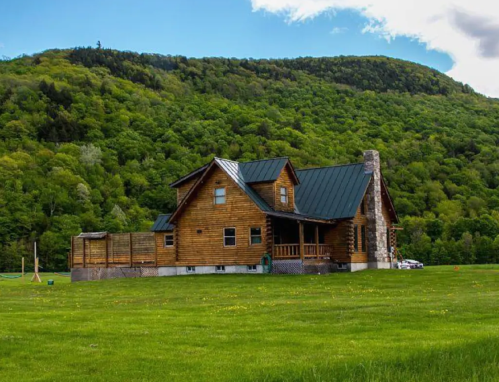 A large wooden house with a green metal roof, set against a backdrop of lush green hills and a clear blue sky.