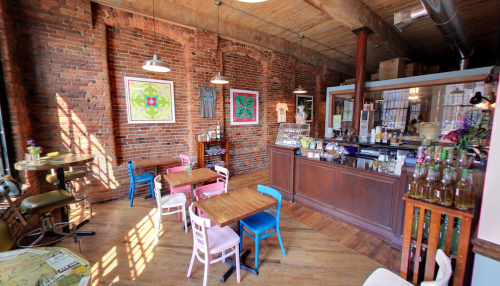 Cozy café interior with brick walls, colorful chairs, wooden tables, and a counter displaying drinks and art on the walls.