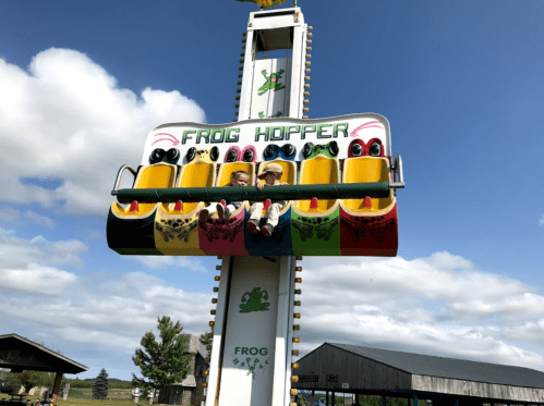 Two children enjoy the Frog Hopper ride, suspended high against a blue sky with fluffy clouds.