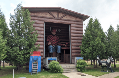 A large statue of Paul Bunyan sits in a log cabin, surrounded by trees and a blue mailbox.