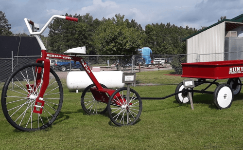 A red tricycle with a wagon attached, set on green grass with trees and a building in the background.
