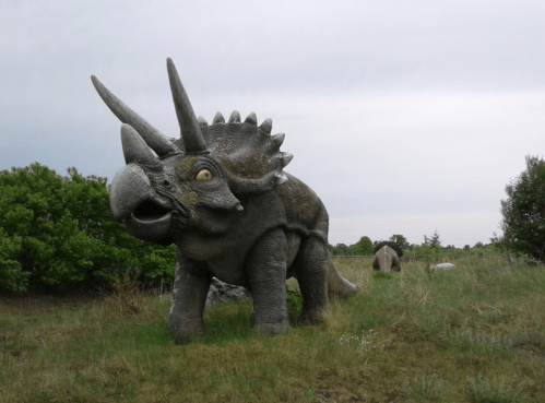 A large, realistic dinosaur statue of a Triceratops in a grassy area with trees in the background.