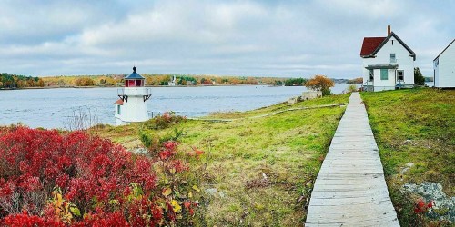 A scenic view of a lighthouse and a house by a river, surrounded by autumn foliage and a wooden walkway.