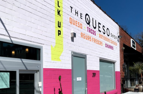 Colorful exterior of The Queso Shop with a pink and white wall, featuring a menu sign and outdoor seating.