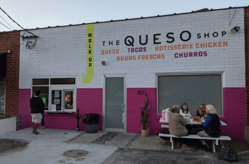 Exterior of The Queso Shop featuring a colorful sign and a group of people dining at a picnic table.