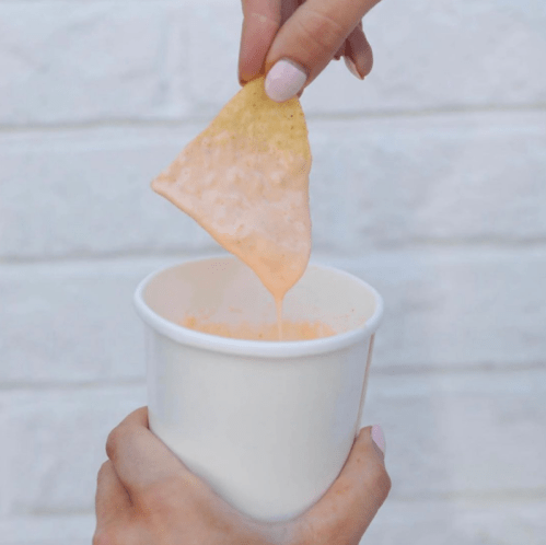 A hand dips a tortilla chip into a cup of creamy orange sauce against a white background.