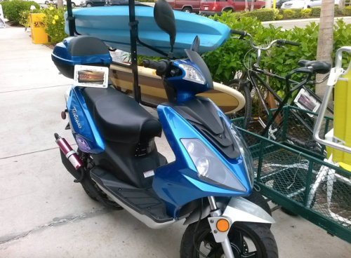 A blue scooter parked on a sidewalk, with a kayak and bicycles in the background.