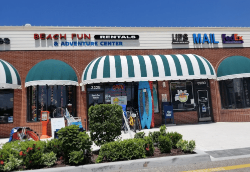 Storefront of Beach Fun Rentals & Adventure Center with colorful surfboards and a UPS/FedEx sign. Bright, sunny day.