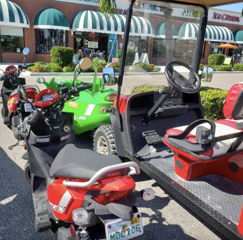 A row of colorful vehicles, including scooters and a golf cart, parked in a sunny outdoor area with shops in the background.