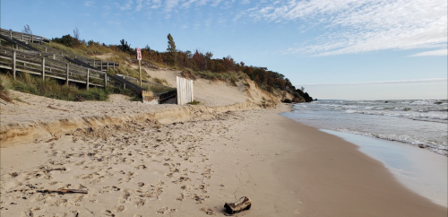 A sandy beach with gentle waves, wooden stairs leading up to dunes, and a clear sky in the background.