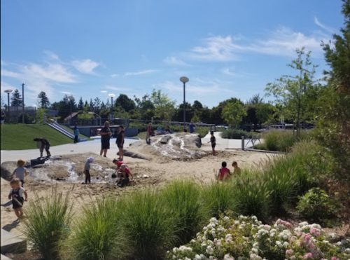Children playing in a sandy area with water features, surrounded by greenery and a clear blue sky.