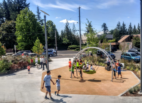 A sunny park scene with children playing on a swing set, surrounded by trees and families enjoying the outdoors.
