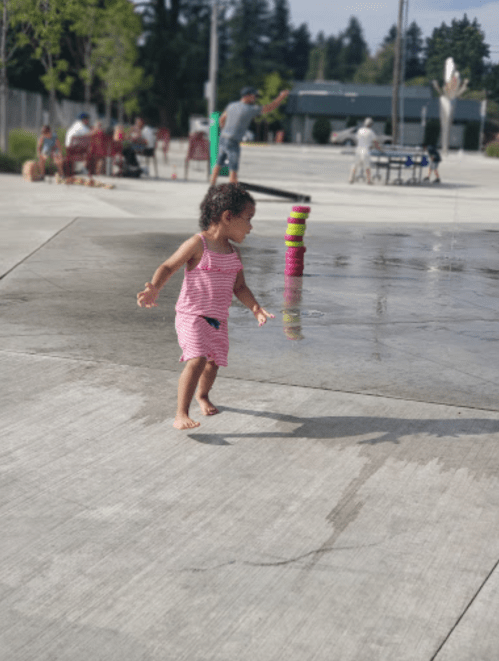A young child in a striped dress plays in a splash pad, with colorful cups stacked nearby and people in the background.