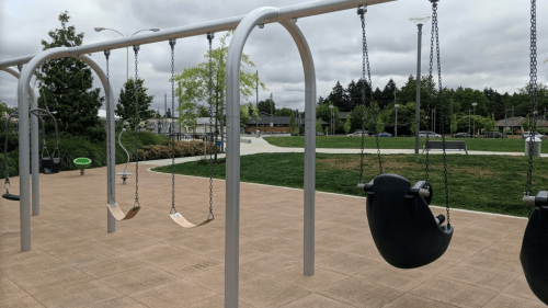 A playground with empty swings hanging from a metal frame, surrounded by green grass and trees under a cloudy sky.
