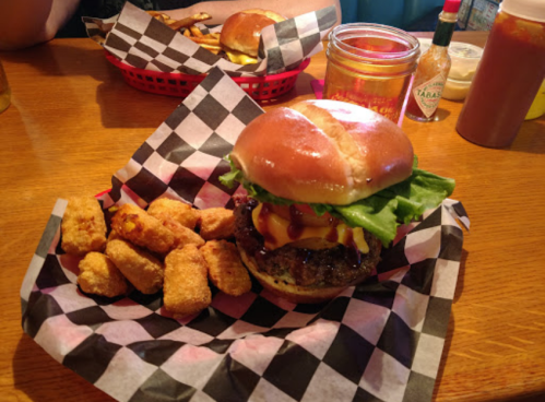 A juicy burger with lettuce, cheese, and condiments, served with crispy fried snacks on a checkered paper plate.